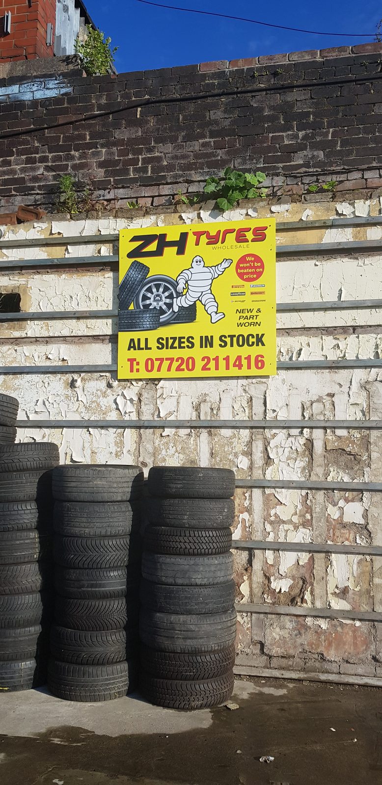 The yellow ZH Tyres Wholesale sign on the yard wall reading all sizes in stock, with the phone number